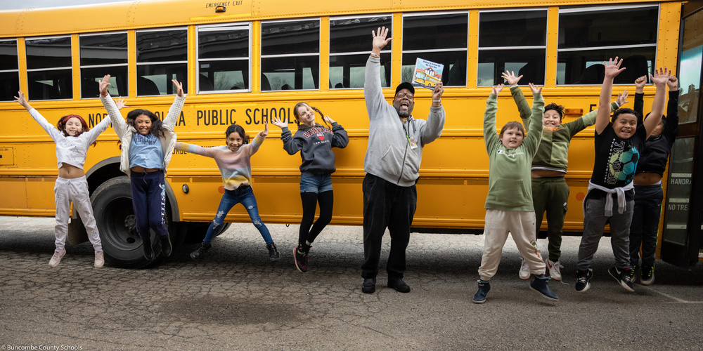 Mr. Cee and several Emma Elementary School students jump for joy in front of a school bus.