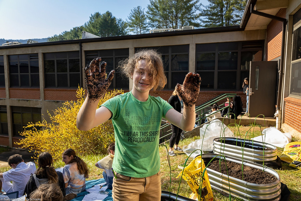 A student holds up dirt-covered hands while standing in front of the garden.
