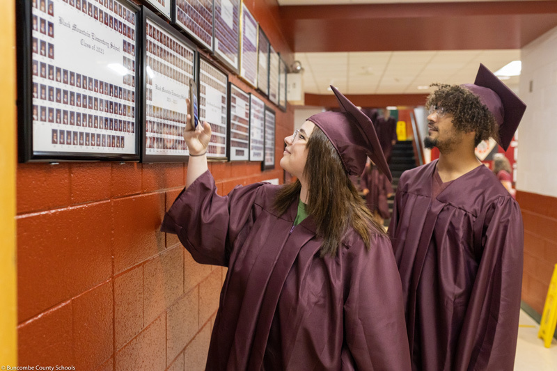 Two seniors in caps and gowns point at old class photos  on the wall of Black Mountain Elementary.