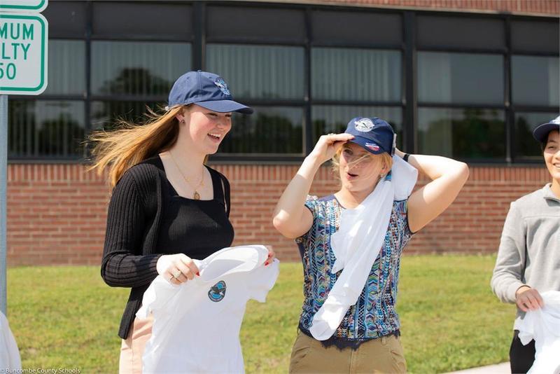 Two students wearing hats and holding t-shirts