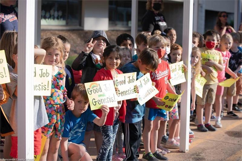 Students lined up in front of a school holding signs that say good job Genesis