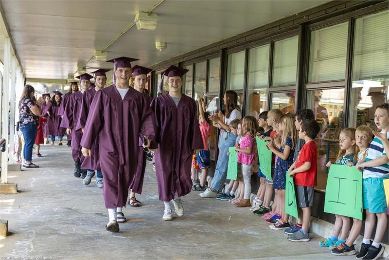Graduates walking in a line down the breezeway with younger students lined up along the wall
