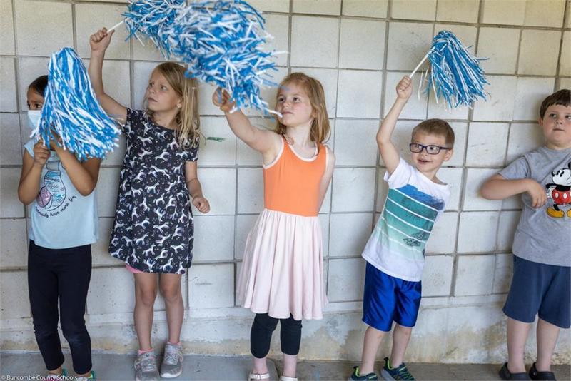 Younger students lined up along the wall holding pompoms