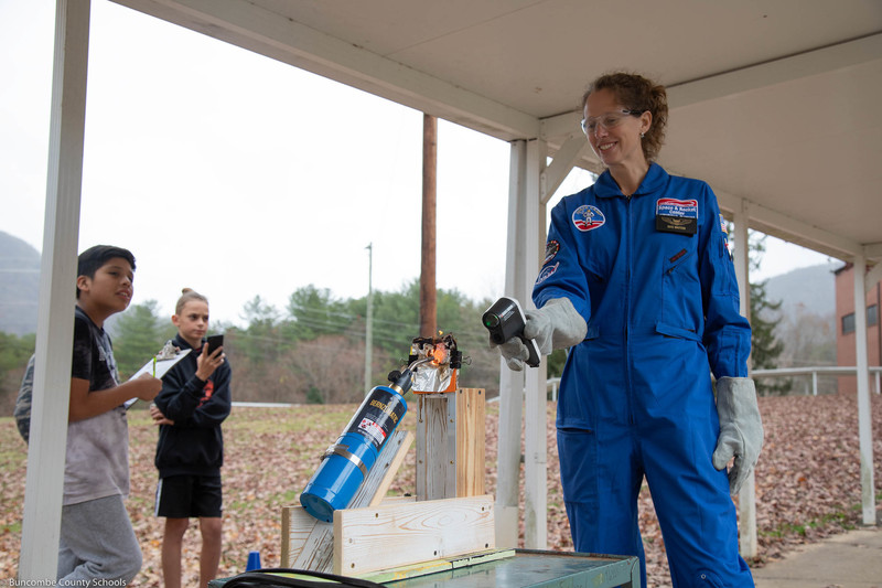 Mrs. Whittier holding a thermometer to test the heat of the egg as the blowtorch works on the heat shield.